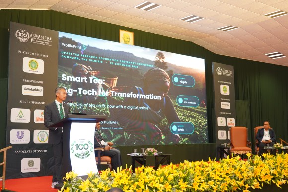 A speaker presenting at the UPLAST Tea Research Foundation Conference, with a large screen displaying a presentation titled 'Smart Tea: Ag-Tech for Transformation' in the background.
