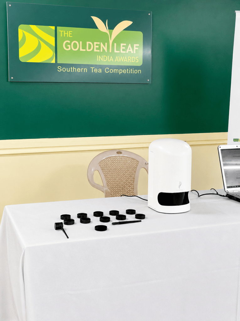 A table set up for the Golden Leaf India Awards Southern Tea Competition, featuring a sign on a green wall, a white device, a laptop, and several black containers.