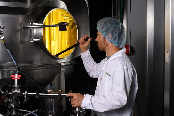A technician in a hairnet and white lab coat is inspecting an industrial machine, examining the interior through a circular opening, with a yellow background visible inside the machine.